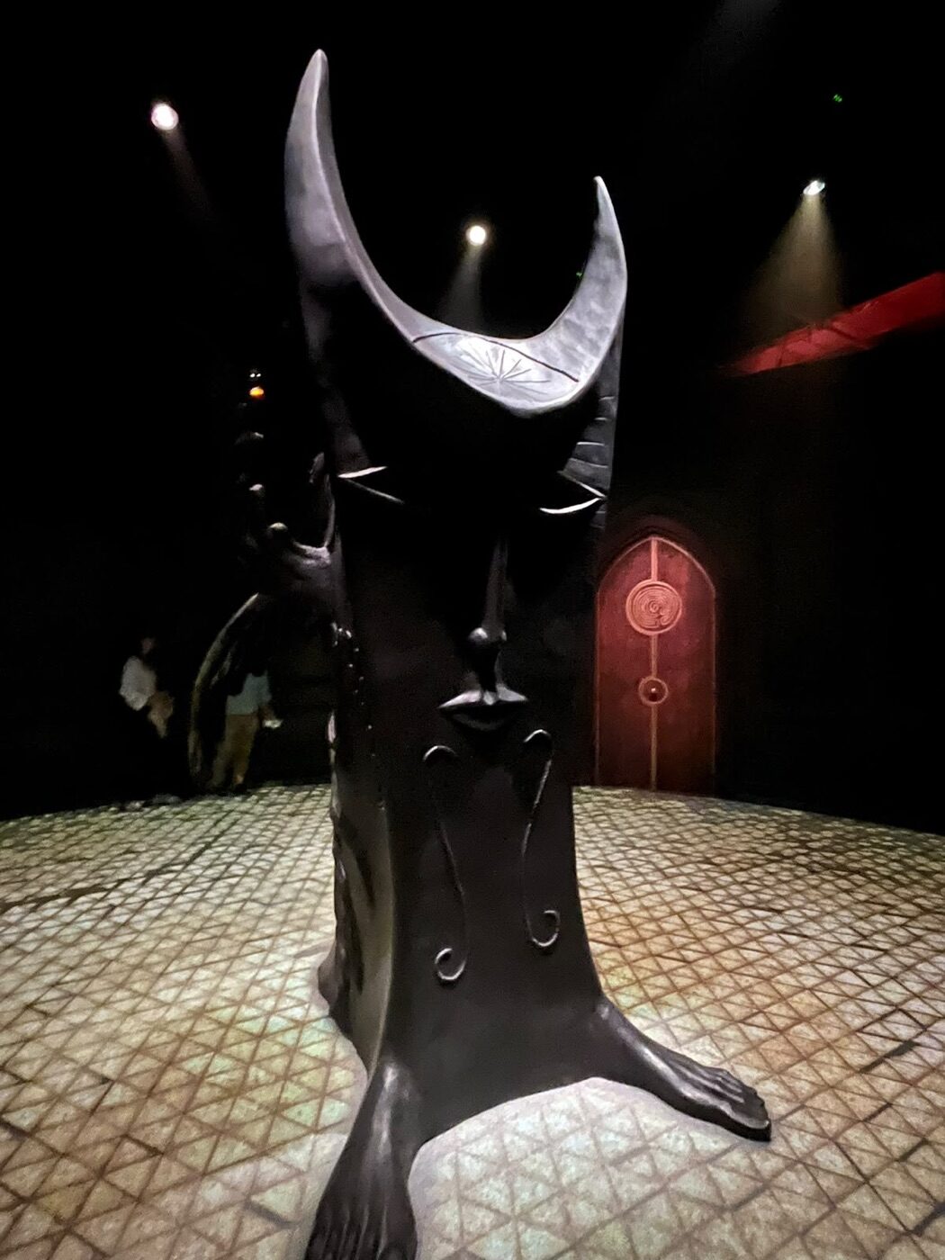 Escultura oscura de Leonora Carrington con tocado de luna creciente sobre piso de mosaico, con una puerta roja con espiral al fondo
