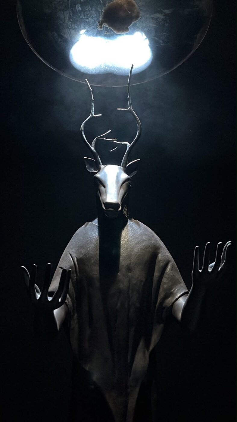 Escultura de figura con cabeza de venado y astas alzando las manos bajo una esfera luminosa en la exposición Laberinto Mágico