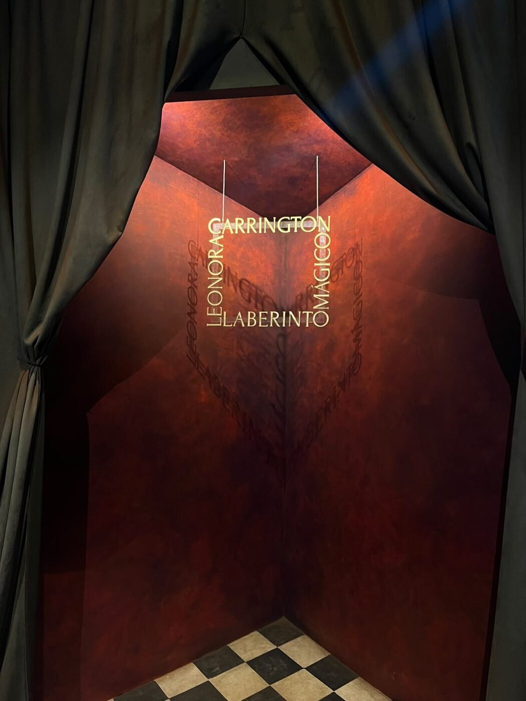 Acceso con cortinas oscuras, muro rojo, letrero de neón de Leonora Carrington Laberinto Mágico y piso de ajedrez blanco y negro