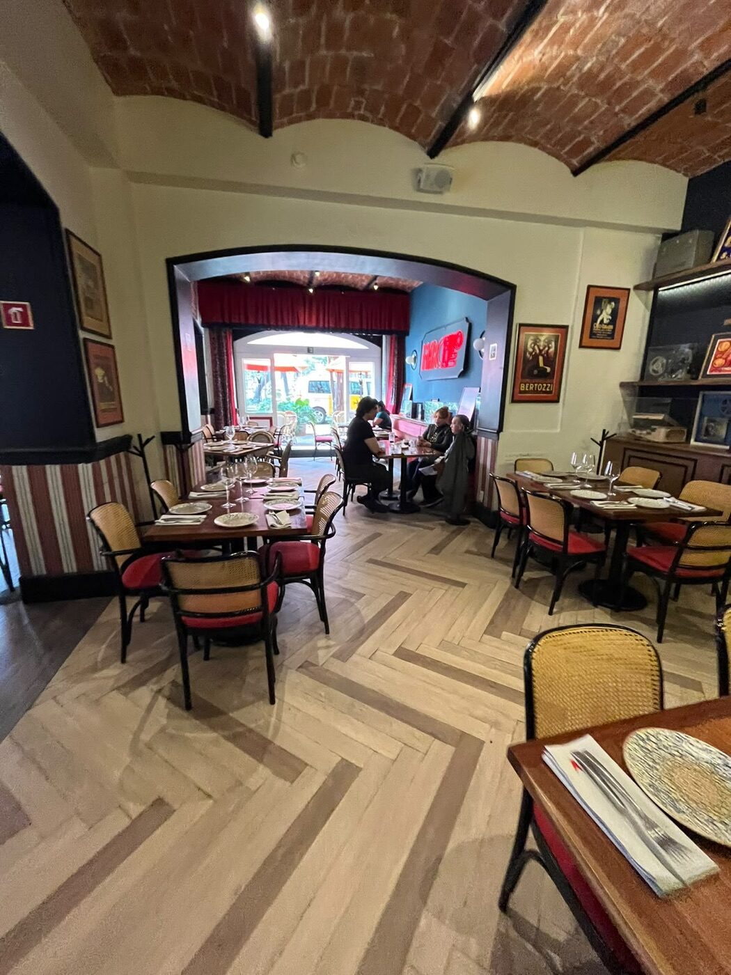 Comedor de Marcello con bóveda de ladrillo, piso de espiga y sillas de rejilla con asiento rojo, y el letrero de neón al fondo