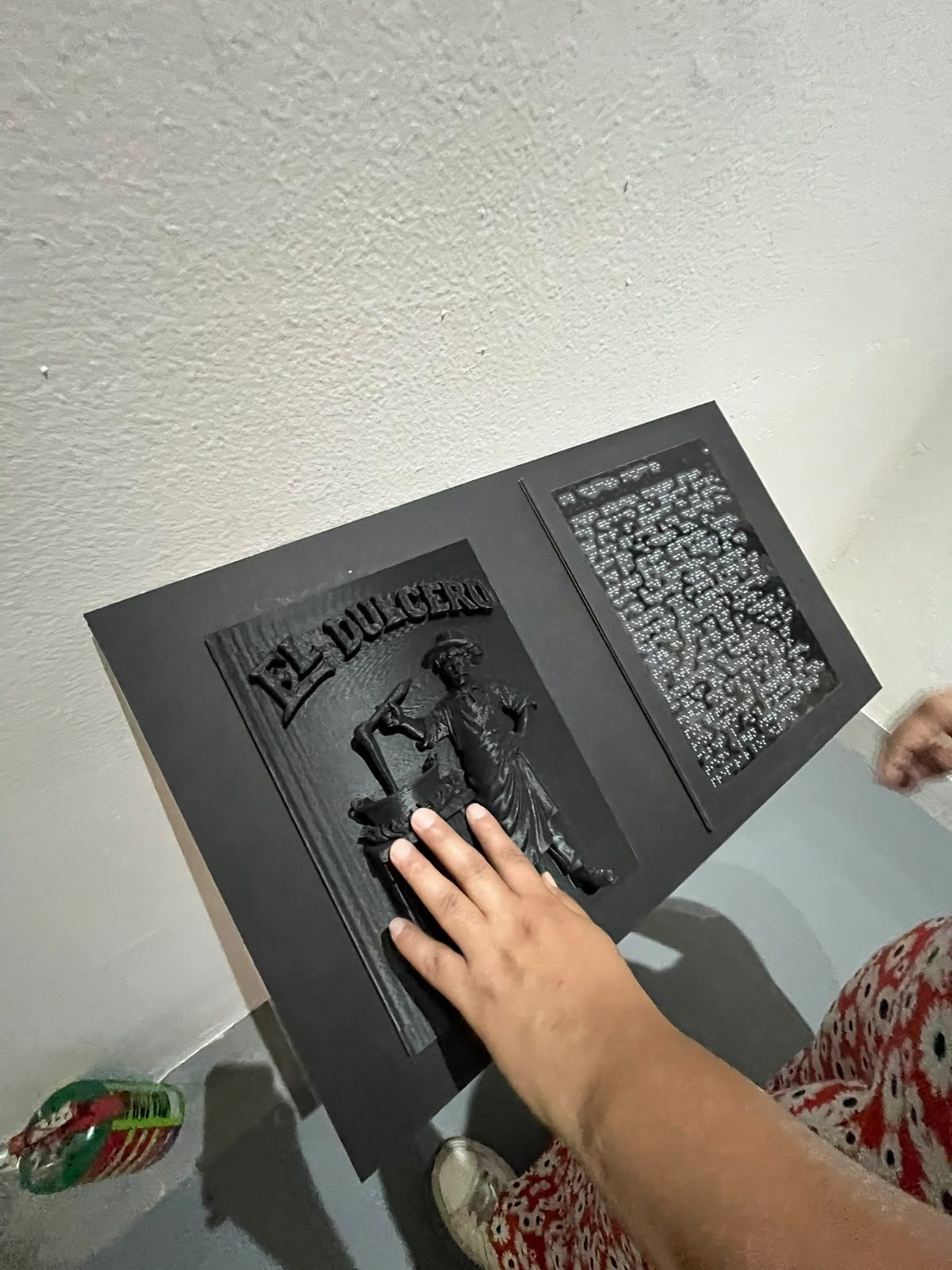 Pieza táctil en relieve de El Dulcero con placa en braille en la exposición de Posada del Munae, con una mano recorriéndola