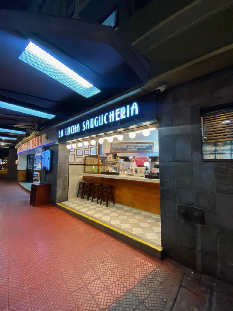 Fachada iluminada de La Lucha Sanguchería en Miraflores, de noche, con barra y bancos de madera, enero de 2025